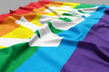 Flagge mit sechs waagerechten Streifen in den Farben Rot, Orange, Gelb, Grün, Blau und Violett. Auf der Flagge steht in weißen Großbuchstaben das Wort 'PACE'. Die Flagge ist leicht gewellt und liegt auf einer Fläche.