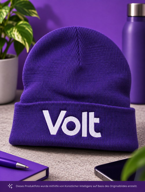 Lila Strickmütze mit weißem Schriftzug 'Volt' auf einem Tisch mit lila Notizbuch, Stift, Smartphone und Flasche im Hintergrund
