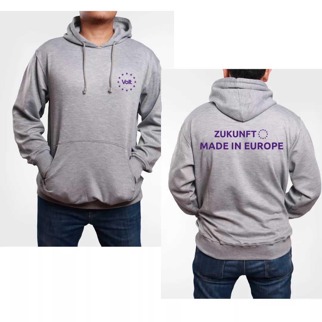 Hellgrauer Kapuzenpullover mit Kängurutasche, vorne links auf der Brust ein violettes Logo mit dem Schriftzug 'Volt' umgeben von zwölf Sternen in Kreisform. Auf der Rückseite in violetter Schrift der Text 'ZUKUNFT MADE IN EUROPE' mit einem Kreis aus zwölf Sternen rechts daneben. Die Person trägt dunkle Jeans, die Kapuze liegt locker auf dem Rücken.