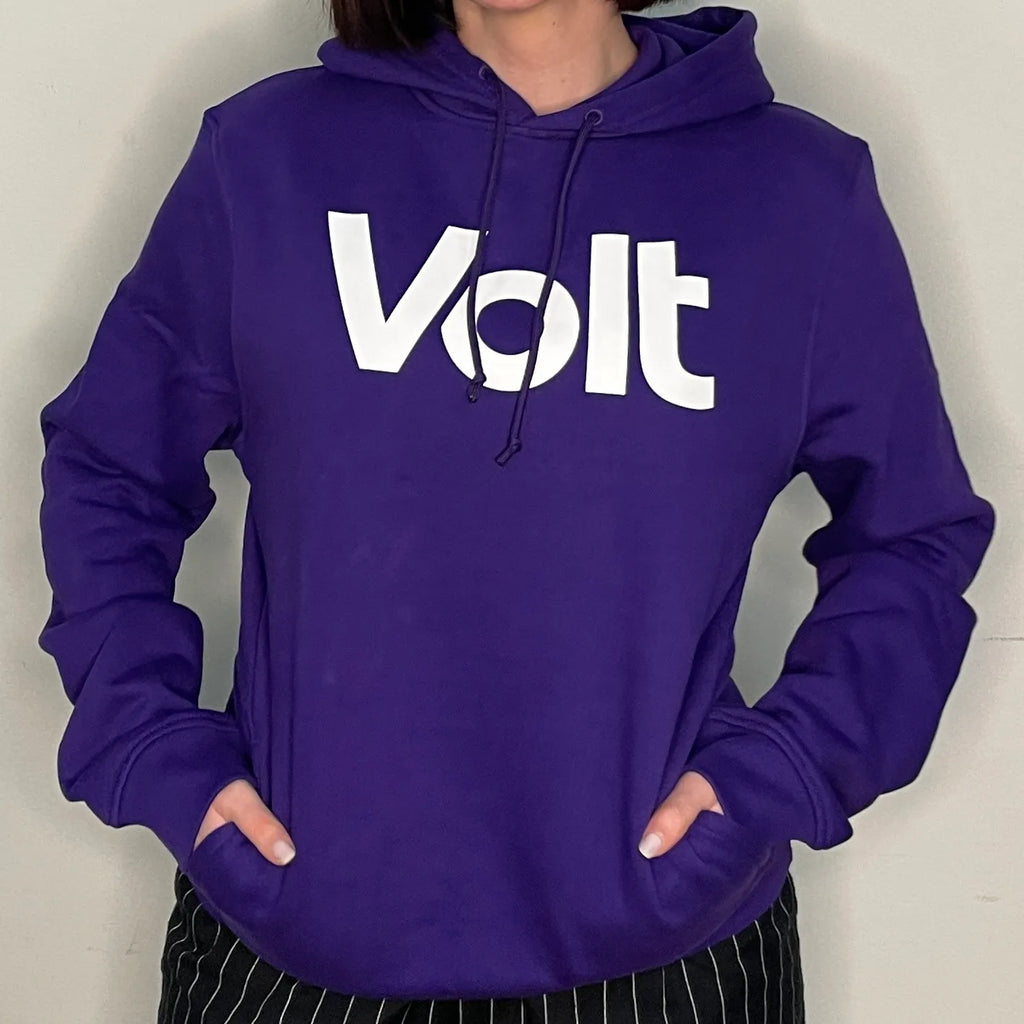 Person trägt einen violetten Kapuzenpullover mit der weißen Aufschrift 'Volt' auf der Brust. Die Hände sind in den Taschen des Pullovers. Die Person trägt eine schwarz-weiß gestreifte Hose.