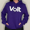 Person trägt einen violetten Kapuzenpullover mit der weißen Aufschrift 'Volt' auf der Brust. Die Hände sind in den Taschen des Pullovers. Die Person trägt eine schwarz-weiß gestreifte Hose.