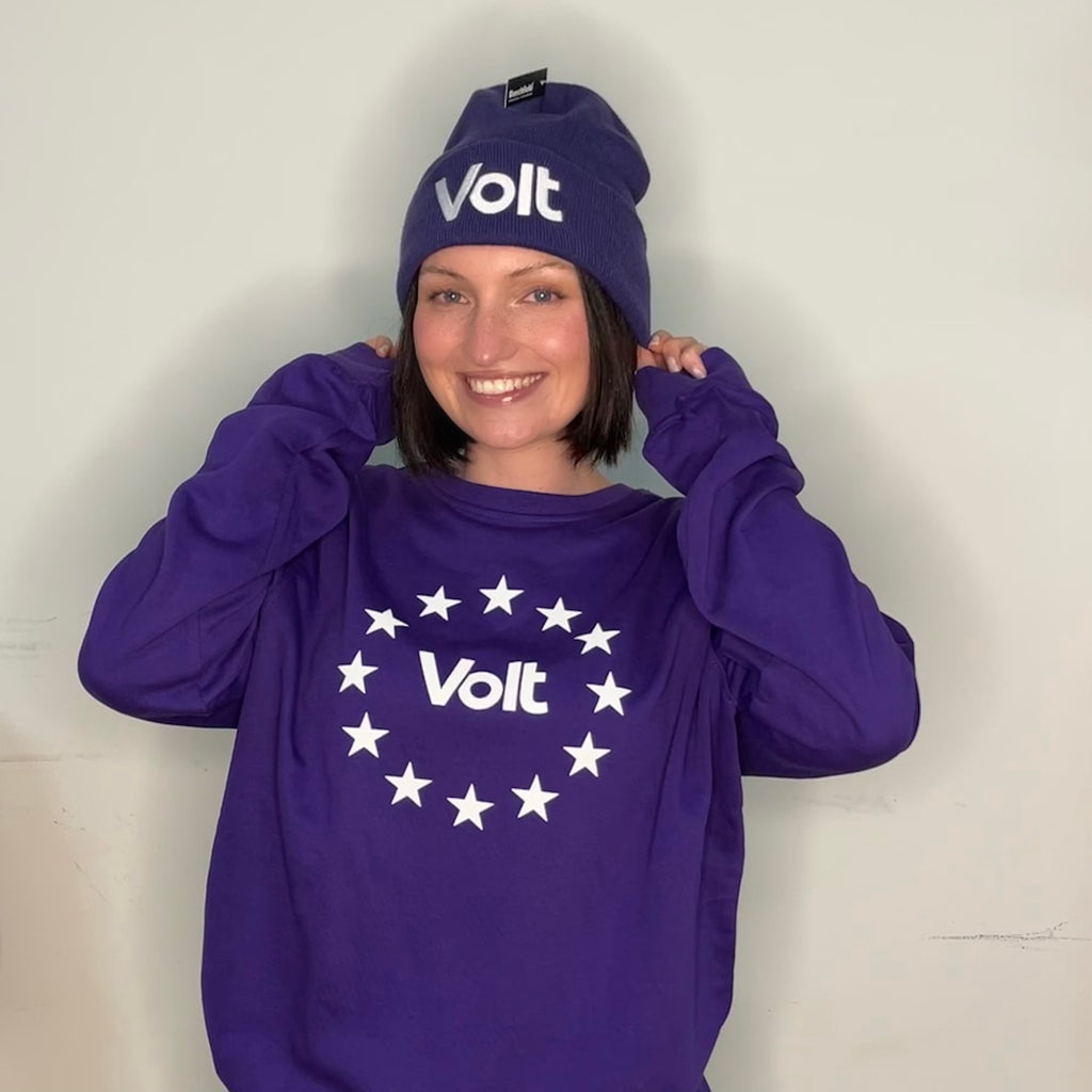 Person trägt eine lila Mütze mit dem weißen Schriftzug 'Volt' und ein lila Sweatshirt mit einem weißen Kreis aus Sternen und dem Schriftzug 'Volt' in der Mitte. Die Person hält mit beiden Händen die Mütze am Kopf.