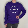 Person trägt ein violettes Sweatshirt mit weißem Schriftzug 'Volt' in der Mitte, umgeben von zwölf weißen Sternen in einem Kreis. Die Person steht mit einer Hand in der Hüfte, trägt dunkle Jeans und ist vor einem hellen Hintergrund zu sehen.