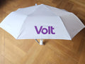 Weißer Taschenregenschirm mit violetter Aufschrift 'Volt' auf einer der Schirmflächen, geöffnet auf einem hellbraunen Parkettboden liegend.