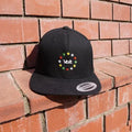 Schwarze Baseballkappe mit flachem Schirm, auf rotem Ziegelsteinmauerstück platziert. Auf der Vorderseite ist ein Kreis aus zwölf kleinen Sternen in verschiedenen Farben (rot, gelb, grün, blau, weiß) gestickt. In der Mitte des Sternenkreises steht in weißer Schrift das Wort 'Volt'. Auf dem Schirm klebt ein silberner Aufkleber mit schwarzer Schrift.