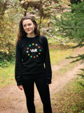 Eine Person steht auf einem Waldweg, umgeben von Bäumen und Sträuchern. Sie trägt ein schwarzes Sweatshirt mit dem Schriftzug 'Volt' in der Mitte, umgeben von einem Kreis aus zwölf Sternen in verschiedenen Farben. Die Person trägt schwarze Hosen und hat lange, gewellte Haare.