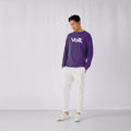 Ein Mann steht vor einer weißen Wand. Er trägt ein lila Sweatshirt mit dem weißen Schriftzug 'Volt' auf der Brust, weiße Hosen und weiße Sneaker. Seine Hände sind in den Hosentaschen. Der Boden ist hell und glatt.